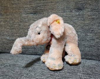 Pode incluir: Um brinquedo de elefante de pelúcia rosa e cinza com um sino no pescoço. O elefante tem uma flor amarela na orelha e uma fita vermelha ao redor do pescoço.