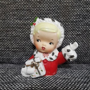 May include: A porcelain figurine of a girl wearing a red dress and a white fur-trimmed hat. She is holding a wrapped gift box with a gold ribbon and holly sprigs.