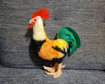 May include: A plush rooster figurine with a red comb and wattle, a yellow and orange body, and a green tail. The rooster has tan feet and is set against a gray textured background.