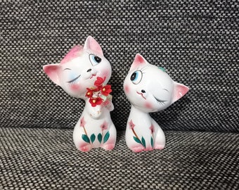 May include: Two white ceramic figurines of cats with pink accents and winking eyes. One cat is holding a bouquet of red flowers. Both cats have green leaves and pink flowers painted on their bodies.