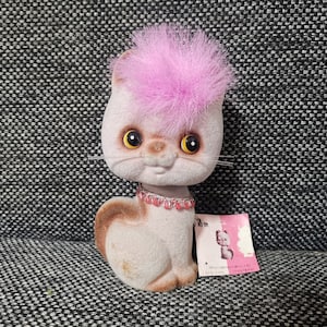 May include: A pink and brown plush cat toy with a pink fur pom-pom on its head. The cat is wearing a pink beaded necklace and has yellow eyes. The toy is sitting on a gray textured surface.