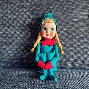 Op de afbeelding: Een vintage elfenpop met blonde vlechten, die een groene outfit met capuchon draagt met rode afwerking en blauwe schoenen. De pop zit op een grijs, getextureerd oppervlak.
