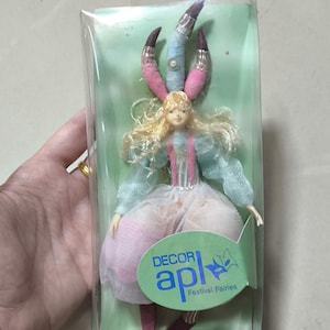Peut inclure: Une poupée de fée dans un emballage plastique transparent. La poupée a des cheveux blonds, une robe rose et bleue, et une coiffe élaborée. L'emballage a un dos vert et une étiquette qui dit "DECOR apl Festival Fairies."