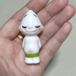 May include: A small, white figurine with a conical head, closed eyes, and rosy cheeks. The figurine has a green band around its waist and is held in a hand. The figurine is a collectible item.