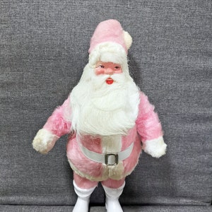 Puede incluir: Una figura de Papá Noel de peluche rosa con barba blanca y botas blancas. Papá Noel lleva un gorro rosa con un pompón blanco.