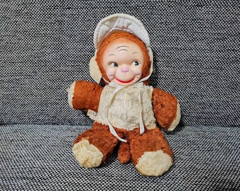 May include: A brown and white plush toy monkey wearing a white bonnet. The monkey has a pink nose and a friendly expression.