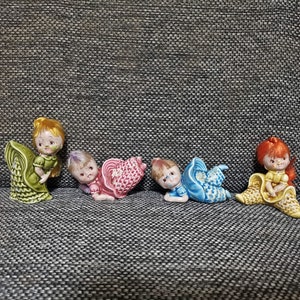 Peut inclure: Cinq figurines en céramique d'enfants vêtus de robes colorées et tenant des coquillages. Les figurines sont disposées en rangée sur une surface texturée grise.