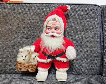 May include: A plush Santa Claus doll wearing a red suit with white trim and a white beard. He is holding a basket with two white plush animals.