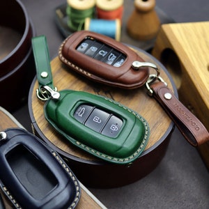 Puede incluir: Tres fundas de cuero para llaves en diferentes colores: marr&oacute;n, verde y azul. Las fundas tienen costuras alrededor de los bordes y est&aacute;n dise&ntilde;adas para ajustarse a los llaveros de los coches. Las fundas de las llaves se muestran sobre una superficie de madera.