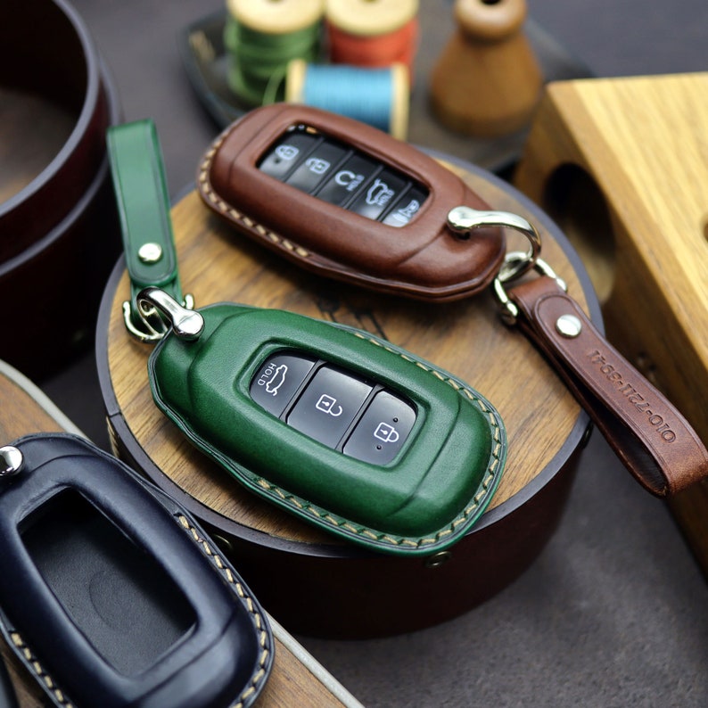 Puede incluir: Tres fundas de cuero para llaves en diferentes colores: marr&oacute;n, verde y azul. Las fundas tienen costuras alrededor de los bordes y est&aacute;n dise&ntilde;adas para ajustarse a los llaveros de los coches. Las fundas de las llaves se muestran sobre una superficie de madera.