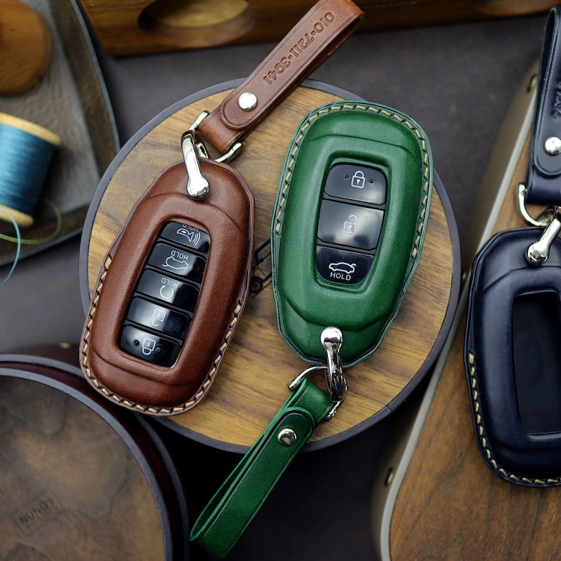 Puede incluir: Dos fundas de cuero para llaves, una marr&oacute;n y otra verde, con detalles de costura. Las fundas tienen varios botones y el texto "HOLD" es visible en la funda verde.
