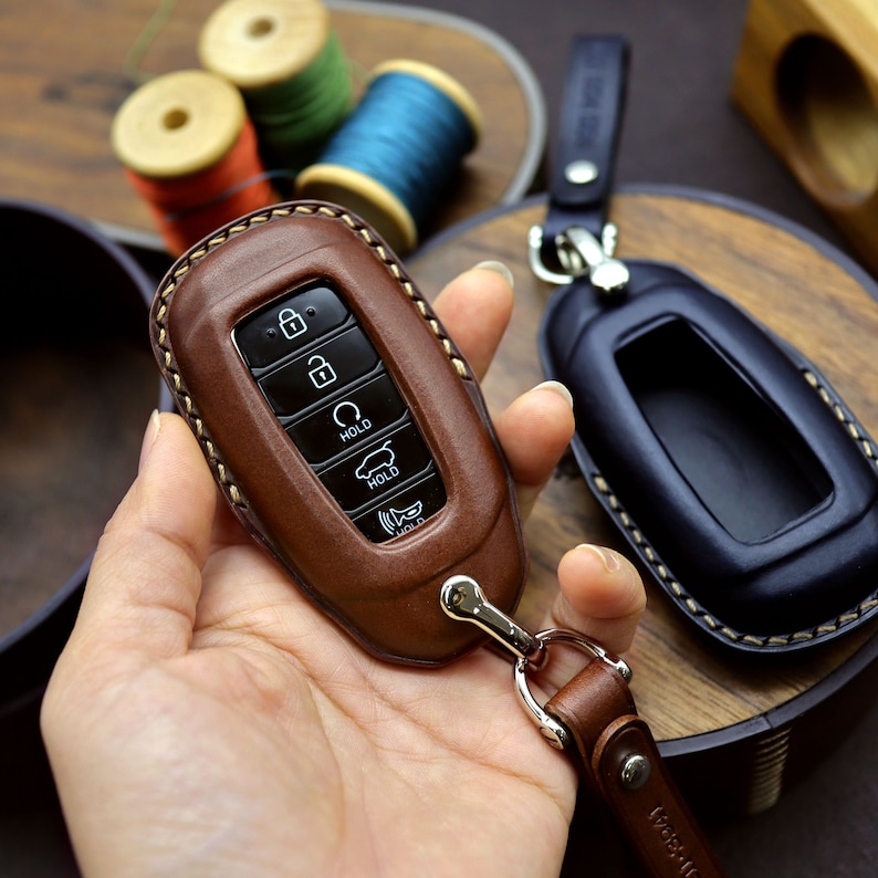 Puede incluir: Un llavero de cuero marr&oacute;n con detalles de costura y un anillo de llave plateado. El llavero tiene varios botones con las palabras "HOLD" y los iconos de un coche, un candado y un altavoz.