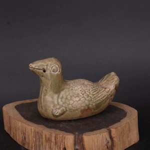 May include: A green glazed ceramic duck figurine with a textured surface. The duck is lying down with its head turned to the side. The figurine is on a wooden base.