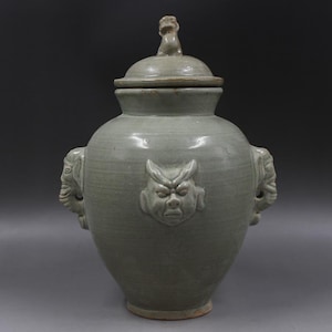 May include: A green glazed ceramic jar with a lid and two elephant-shaped handles. The jar has a carved face on the front.