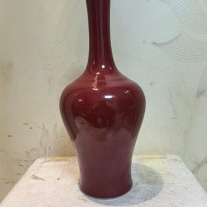 Könnte beinhalten: Eine hohe, glänzende, burgunderfarbene Vase mit schmalem Hals und rundem Körper. Die Vase hat eine glatte, reflektierende Oberfläche und steht auf einer weißen Oberfläche. Die Vase ist ein dekorativer Gegenstand.