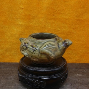 May include: A small, green ceramic pot shaped like a frog with a glazed finish. The pot is sitting on a wooden stand.