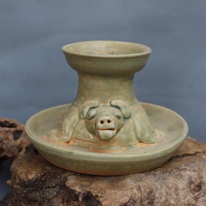 May include: A green ceramic incense burner shaped like a pig. The pig is lying on its back with its legs in the air. The incense burner has a round top and a wide base.