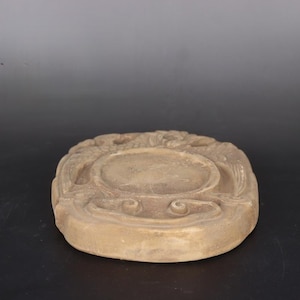 May include: A round, light brown, ceramic incense burner with intricate carvings and a raised center. The burner has a slightly oval shape and a smooth, worn surface.