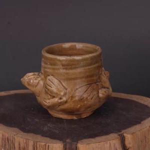 May include: A brown ceramic cup with a frog design. The cup is glazed and has a rustic look.
