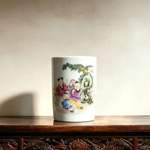 May include: A cylindrical white ceramic vase featuring a colorful hand-painted scene of three children playing outdoors. The artwork depicts figures in traditional clothing near a green rock formation and a tree, rendered in shades of pink, blue, and yellow.