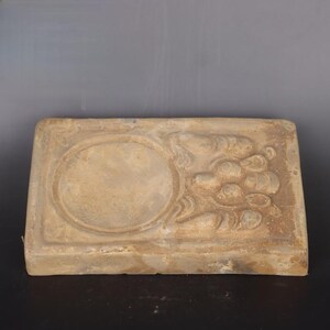 May include: A rectangular, light brown, ceramic tile with a raised, circular design in the center. The tile has a decorative, floral pattern around the circular design.