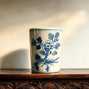 May include: A cylindrical ceramic vase with a cream-colored base and a fine crackle glaze finish. The exterior features a hand-painted blue design depicting a bird perched on a branch beneath a large blooming flower, with foliage surrounding it.