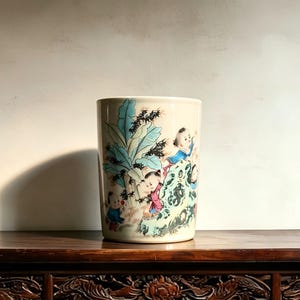 May include: A cylindrical ceramic vessel with an off-white base color, featuring a hand-painted scene of children playing near a large green banana tree and a textured rock formation, rendered in shades of blue, pink, and black ink details.