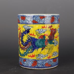 Op de afbeelding: Een cilindrische keramische container met een gele achtergrond en een gedetailleerd feniksontwerp. De feniks is afgebeeld in rood, blauw en groen, met witte wolken. De boven- en onderranden hebben een blauw-wit patroon met rode bloemaccenten.