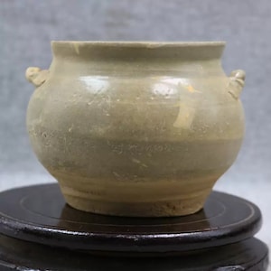 May include: A light brown ceramic pot with two small handles on the sides. The pot has a rounded body and a slightly flared rim.