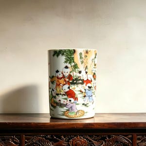 May include: A cylindrical ceramic vessel featuring a colorful hand-painted scene of multiple children playing in a garden setting with green bamboo, rocks, and foliage. The children are depicted wearing traditional clothing in red, yellow, and blue.