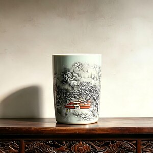 May include: A cylindrical ceramic vase with a pale celadon green base color. The exterior features a detailed monochrome landscape illustration of snow-covered mountains and trees, accented by a single building with a vibrant orange roof.