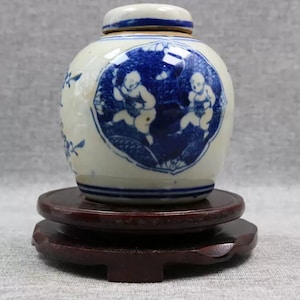 May include: A small blue and white porcelain jar with a lid, decorated with a scene of two children playing. The jar is sitting on a wooden stand.