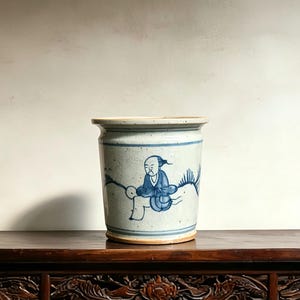 May include: A cylindrical ceramic pot with a speckled off-white glaze and blue hand-painted illustrations. The design features a central figure in blue robes surrounded by stylized foliage, framed by horizontal blue bands at the top and bottom.