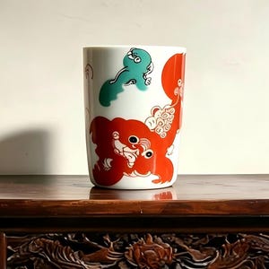 May include: A white ceramic tumbler featuring a vibrant graphic design of a large, stylized red creature with detailed facial features and a smaller, rounded teal figure positioned above it, both rendered in a bold, flat illustrative style.