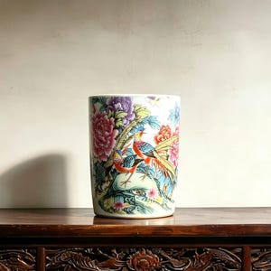 May include: A cylindrical ceramic vase featuring a colorful hand-painted design of two birds perched on a rock surrounded by vibrant pink, purple, and orange peony flowers and green foliage against a white background with a smooth finish.