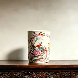 May include: A cylindrical ceramic vase with a cream-colored base featuring a colorful hand-painted design. The artwork depicts a vibrant phoenix with red, blue, and green plumage perched near a large pink peony flower and a flowering tree branch.
