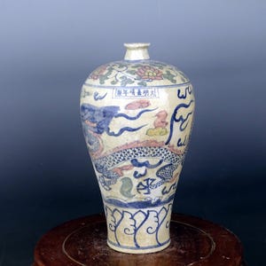 Puede incluir: Un jarrón de cerámica antiguo con base crema, adornado con diseños de dragones y flores azules y rosas. El jarrón presenta caracteres chinos y se exhibe sobre un soporte de madera.