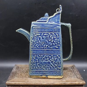 Puede incluir: Una tetera de cerámica azul y blanca con cuerpo cuadrado y tapa. La tetera está decorada con diseños florales y animales en relieve, incluyendo un pájaro y un ciervo. La tetera tiene un pico y una asa.