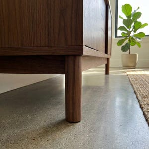 May include: Low-angle view of a wooden cabinet with a rich, dark brown finish. The cabinet has a rectangular shape with visible wood grain and rounded legs. The legs are cylindrical and appear sturdy. The cabinet is set on a concrete floor.
