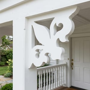 Op de afbeelding: Een close-up van een witte, decoratieve houten beugel met een gestileerd bloemmotief. De beugel is gemonteerd op een witte veranda kolom en overstek. Het ontwerp bevat een hartvorm en bladvormige elementen. Witte deur en balustrade op de achtergrond.