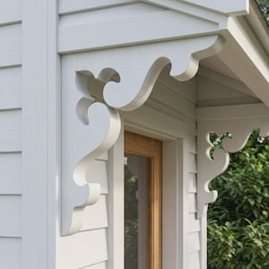May include: A white wooden architectural bracket with a decorative scrollwork design mounted under the eave of a building. The bracket features curved cutouts and a smooth finish, positioned above a doorway with light-coloured horizontal siding.
