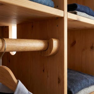 May include: A light-colored wooden closet interior with shelves and a hanging rod. The shelves hold folded clothing. The rod is supported by curved wooden brackets. A wooden hanger with a shirt hangs from the rod.