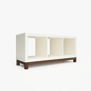 May include: White bookshelf with three open cubbies and a dark wood base. The bookshelf is standing on a white surface.