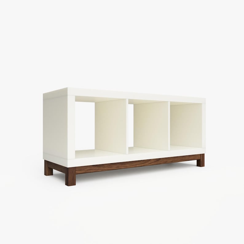 May include: White bookshelf with three open cubbies and a dark wood base. The bookshelf is standing on a white surface.