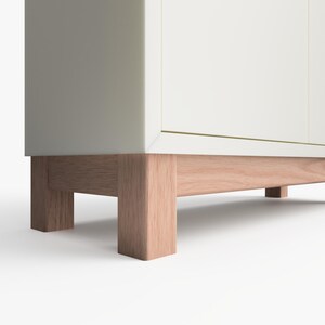 May include: White cabinet with wooden legs. The cabinet has a simple design and is made of a light-colored wood.
