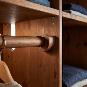 Puede incluir: Primer plano del interior de un armario de madera con estantes y una barra para colgar. La barra está soportada por soportes de madera curvados. Ropa doblada visible en los estantes. La madera tiene un acabado cálido y natural.