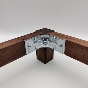 May include: Close-up of a corner joint for a wooden furniture piece. The joint is secured with a metal bracket and a screw. The wood is a dark brown color.