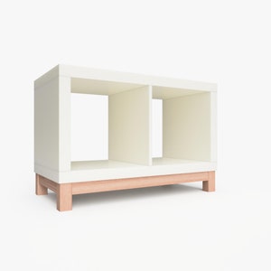 May include: White shelf unit with two open compartments and a natural wood base. The shelf unit is made of wood and has a modern design.