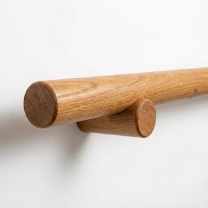 Könnte beinhalten: Ein hölzerner Handlauf mit zylindrischem Design ist an einer weißen Wand befestigt. Der Handlauf besteht aus hellbraunem Holz und wird von einem kleineren, passenden Holzstück getragen. Die Holzmaserung ist sichtbar.