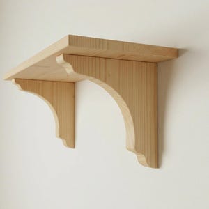 May include: A light-coloured wooden shelf with two decorative brackets. The shelf is rectangular, and the brackets have a curved design. The shelf is mounted on a white wall, showcasing a simple, natural aesthetic.